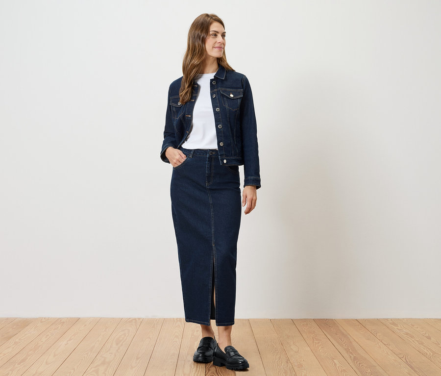 Egy nő áll, farmerkabátot és sliccelt farmer szoknyát visel.