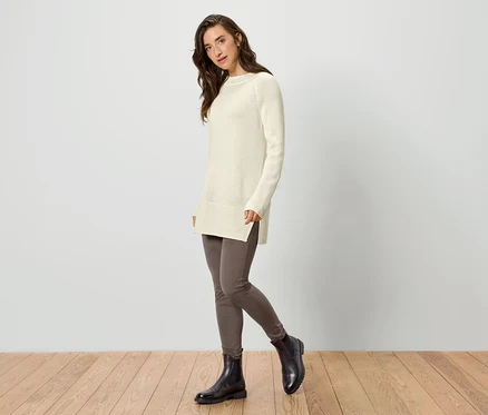 Nő visel krémszínű hosszú kötött pulóvert, leggingset és bokacsizmát.