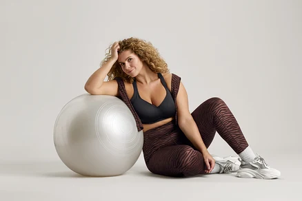 Göndör hajú nő ül egy ezüstszínű gimnasztikai labda mellett, fekete sportmelltartót, zebracsíkos leggingset és tornacipőt visel.