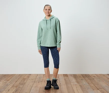 Nő áll világoszöld kapucnis pulóverben, mintás kék Capri leggingsben és fekete túrabakancsban.