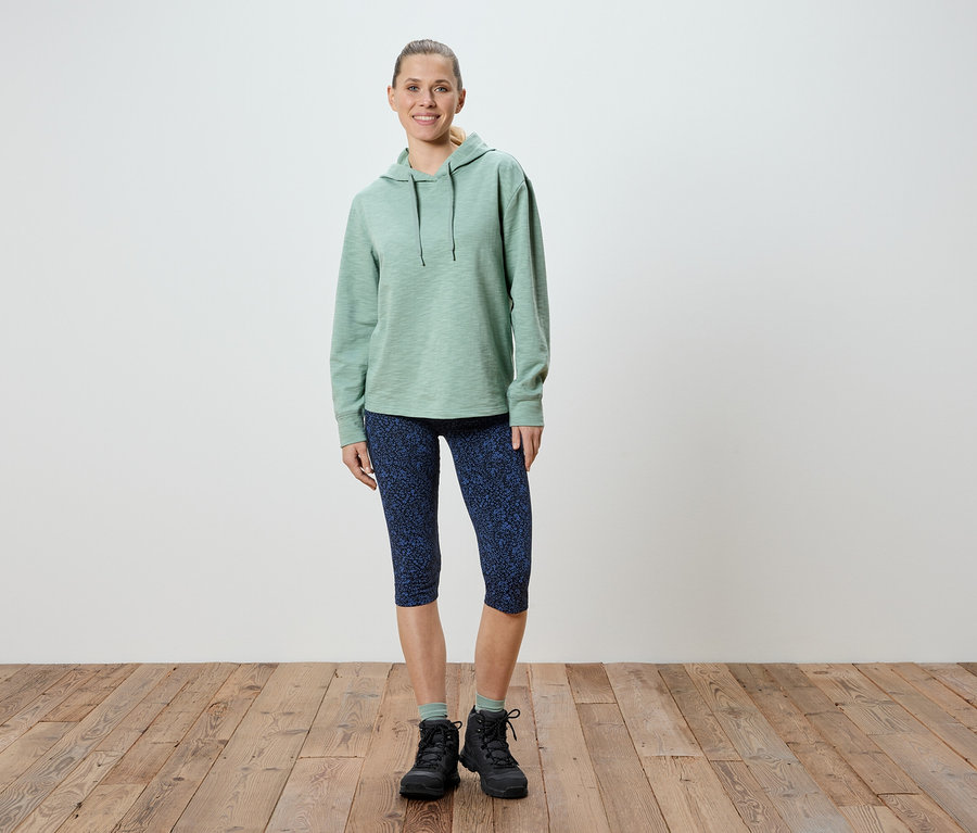 Nő áll világoszöld kapucnis pulóverben, mintás kék Capri leggingsben és fekete túrabakancsban.