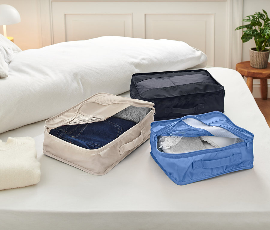 Három nyitott csomagoló kocka fekszik egy ágyon fehér ágyneművel. A kockák bézs, kék és fekete színűek, és ruhákat tartalmaznak.