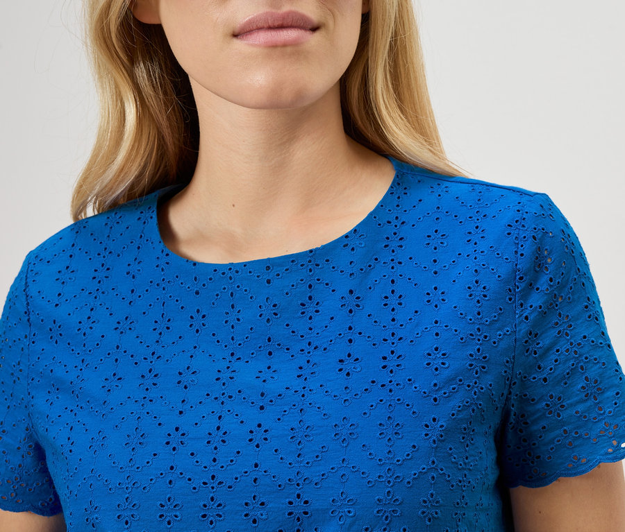 Közeli kép egy kék, angol hímzésű blúzt viselő nőről.