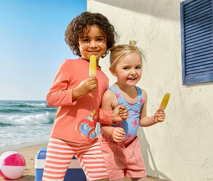 Két gyerek fagylaltozik a strandon. A lány Kinder-Badeanzug, Seesterne fürdőruhát és Kinder-Jeansshorts rövidnadrágot visel. A fiú Kinder-Kombi mit UV-Schutzfaktor 50+, Krake szettet visel.