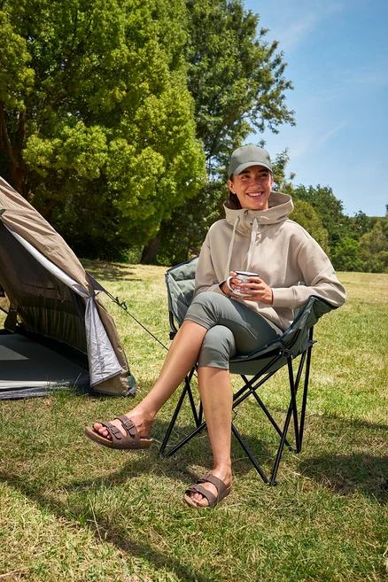 Egy nő ül egy Tambu »ASANA« kempingszéken, egy csészét tart a kezében, és egy 3/4-es outdoor leggingset és egy kapucnis pulóvert visel.