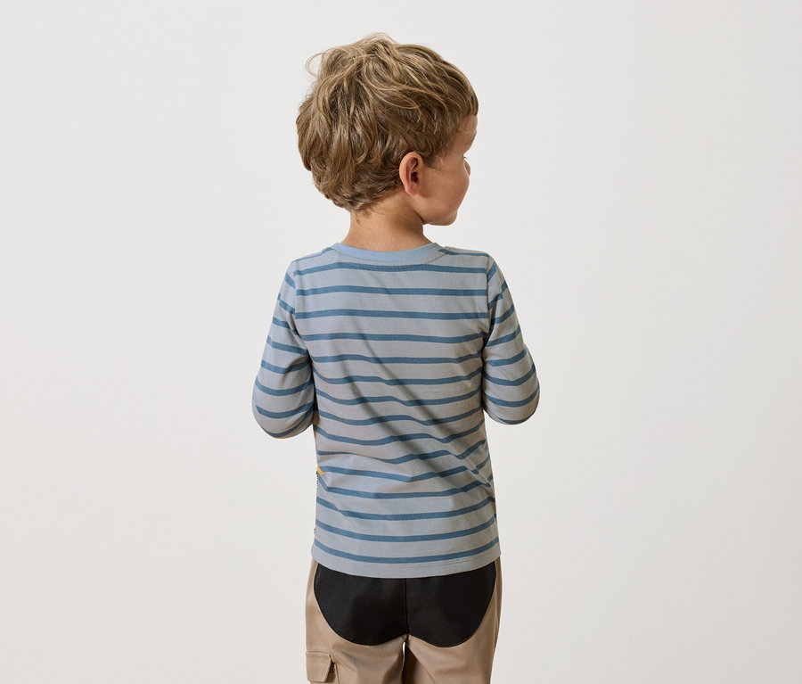 Hátulnézet egy szőke, göndör hajú fiúról, aki kék-szürke csíkos hosszú ujjú inget és bézs nadrágot visel.