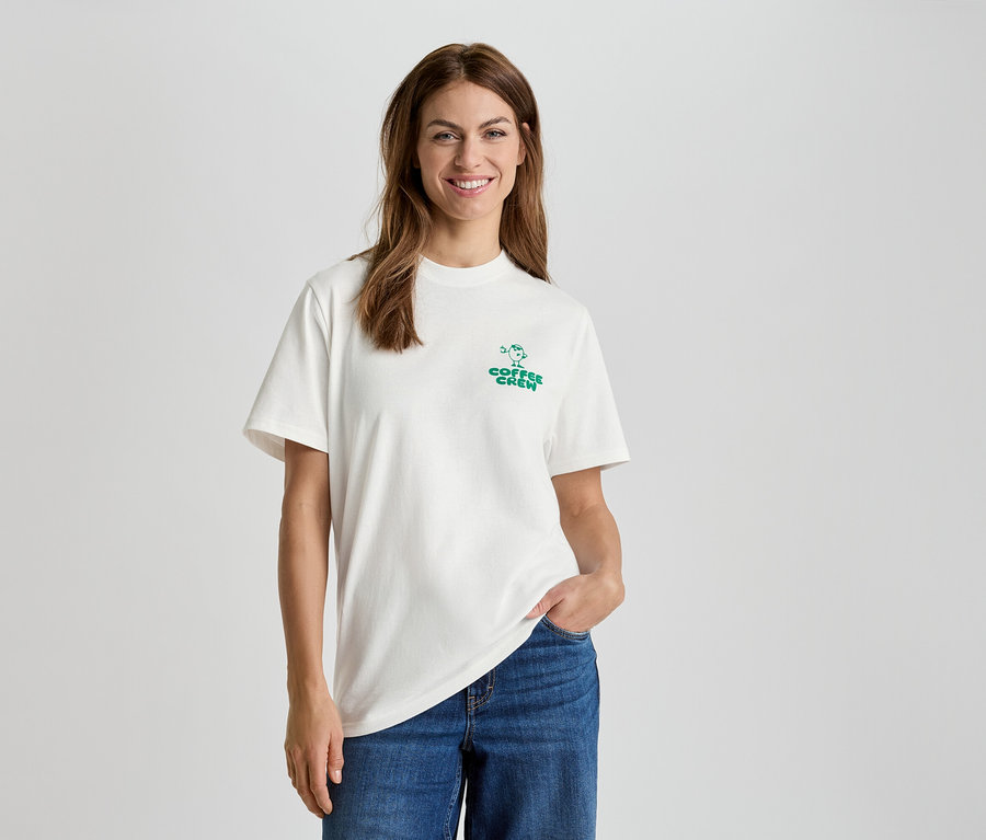 Nő fehér pólóban, "Coffee Crew" felirattal és kék farmerben.