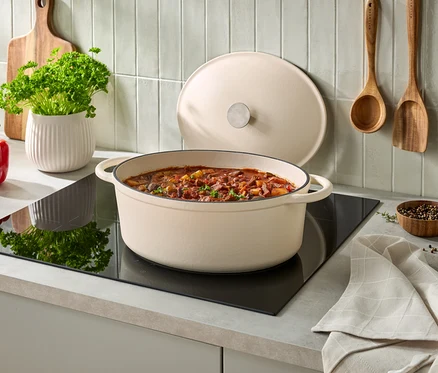 Krém színű ovális öntöttvas edény pörkölttel egy fekete indukciós főzőlapon, fedele mögötte támaszkodik. Balra egy piros kaliforniai paprika, egy fehér cserép petrezselyemmel és egy fa vágódeszka. Két fa konyhai eszköz lóg a csempézett falon, jobbra egy kis tál borssal és egy összehajtott konyharuha.