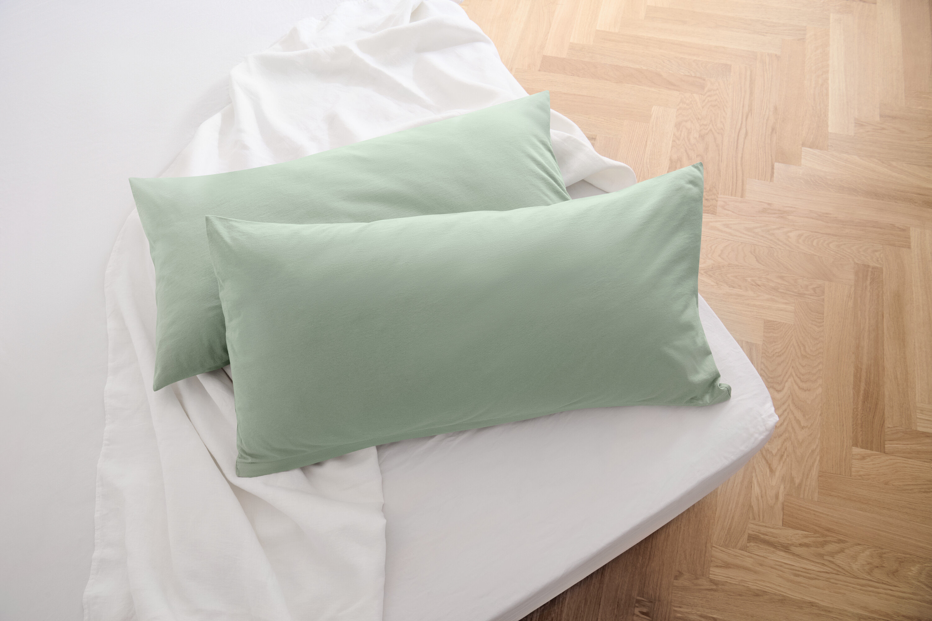 Két zsályaszínű jersey párnahuzat fekszik egy fehér lepedőn.