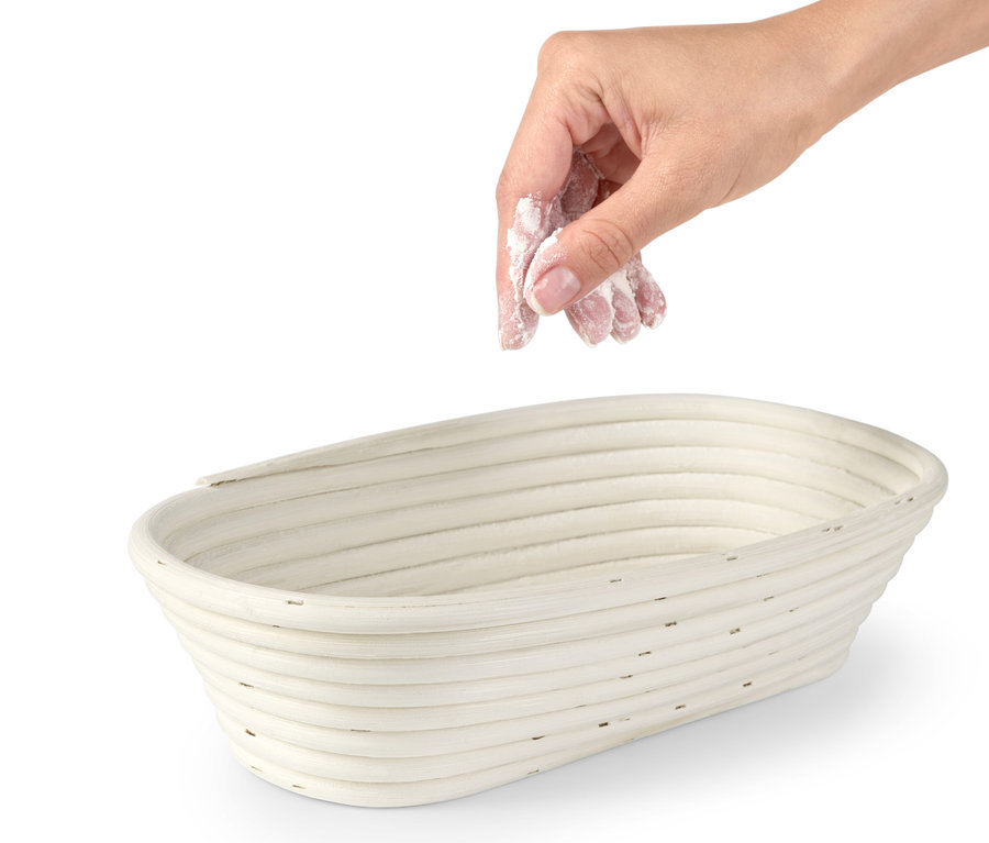 Egy kéz lisztet szór egy ovális kenyérkelesztő kosárba.