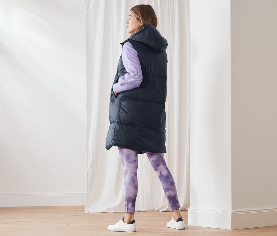 Egy nő áll, és hosszú steppelt mellényt, lila pulóvert és lila leggingset visel.