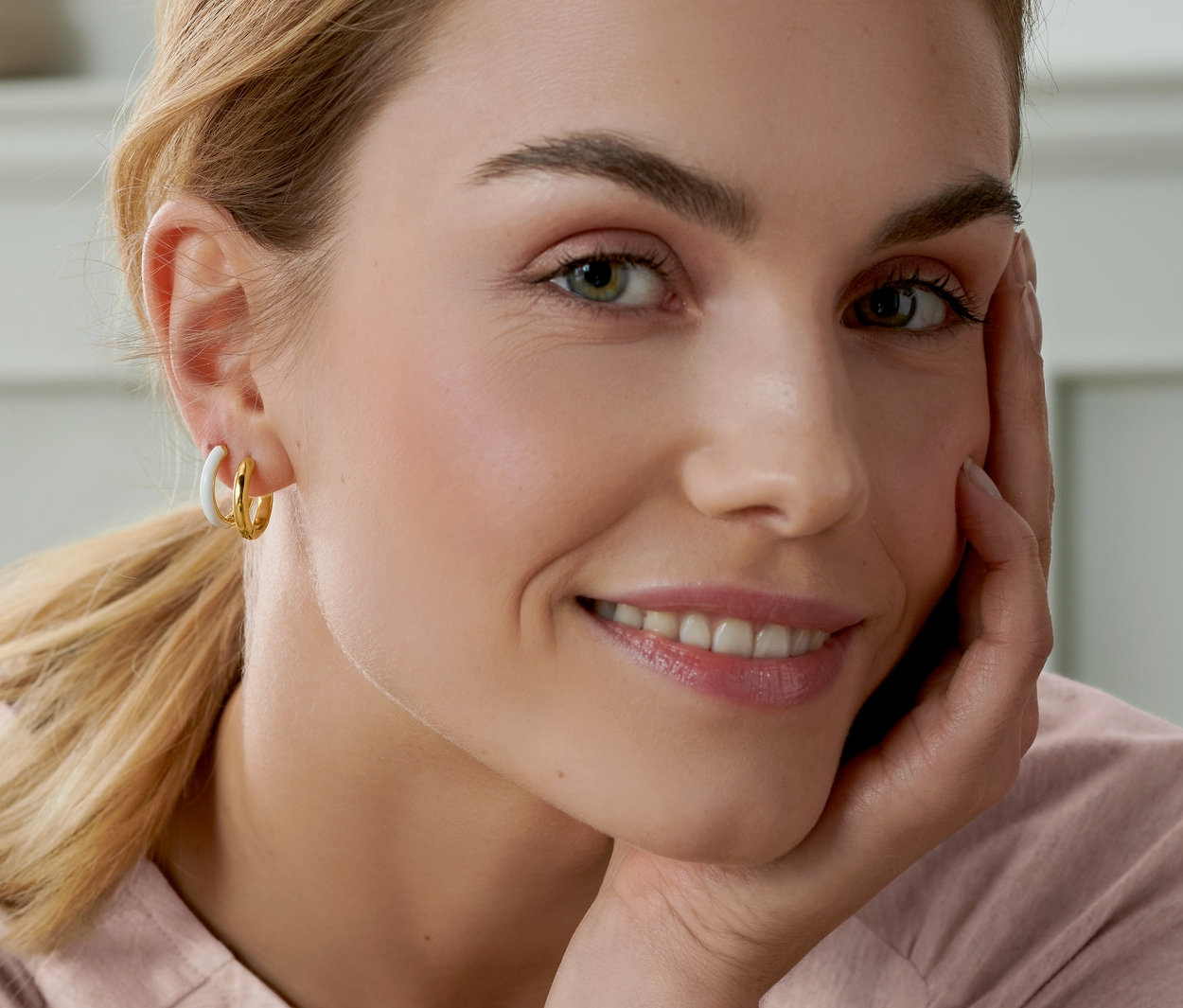 Mosolygó nő az arcát a kezére támasztva, 2 pár aranyozott karika fülbevalót visel.