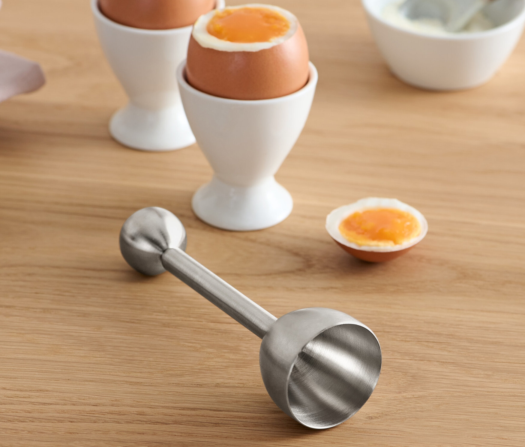 Egy nemesacél tojáshéj törő egy fa asztalon fekszik tojások mellett tojástartóban.