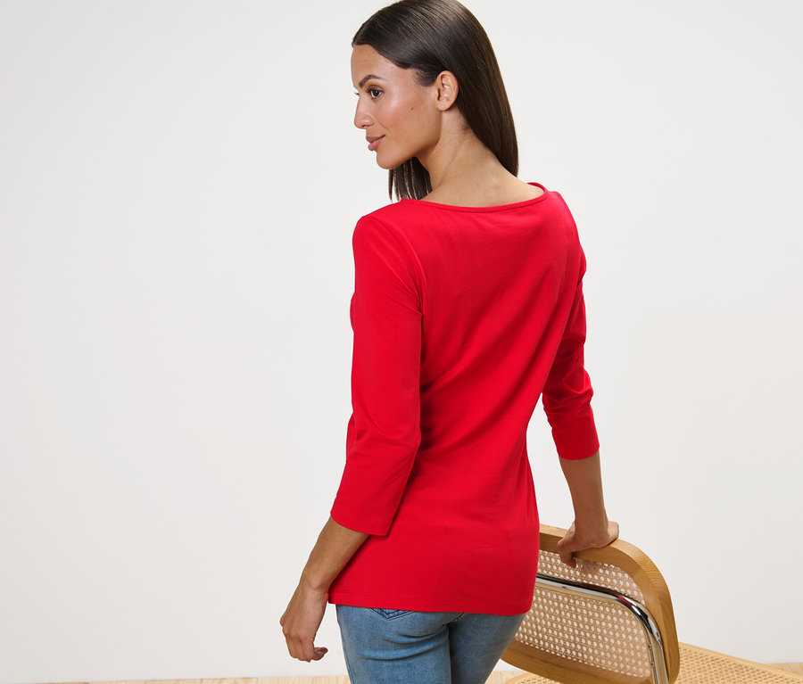 Egy nő a válla fölött néz hátra, és egy székre támaszkodik, piros, háromnegyedes ujjú felsőt visel.