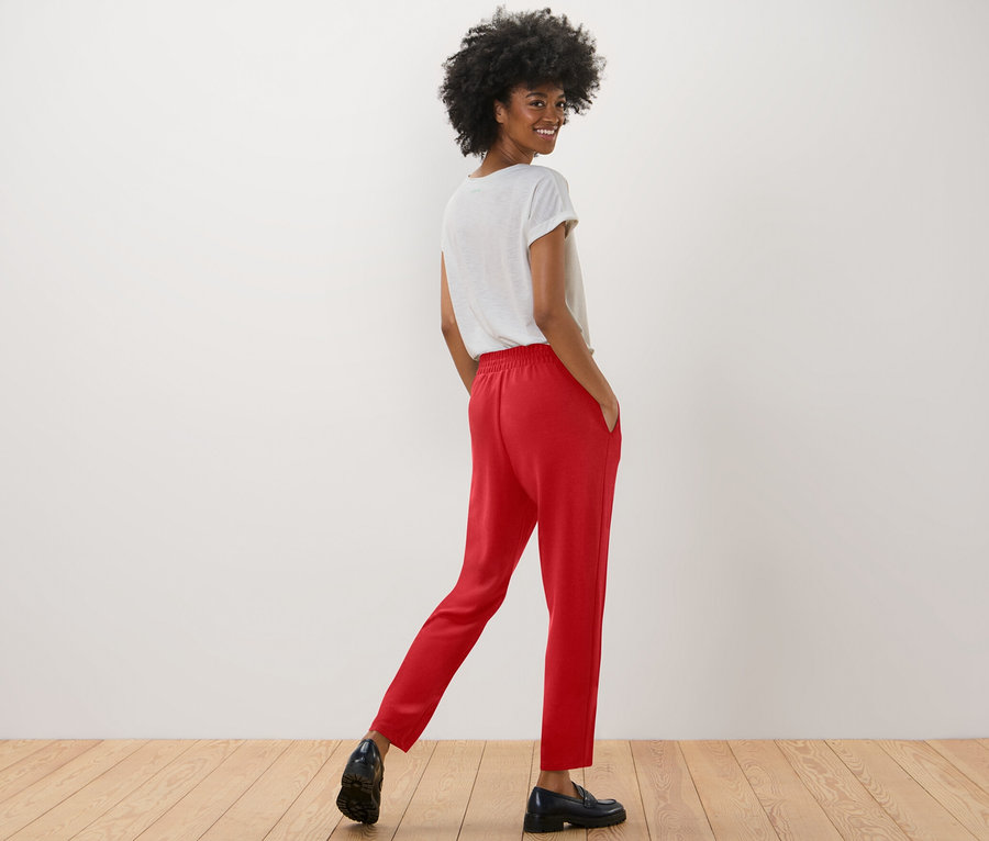 Mosolygó nő pózol piros női lezser nadrágban és fehér pólóban.