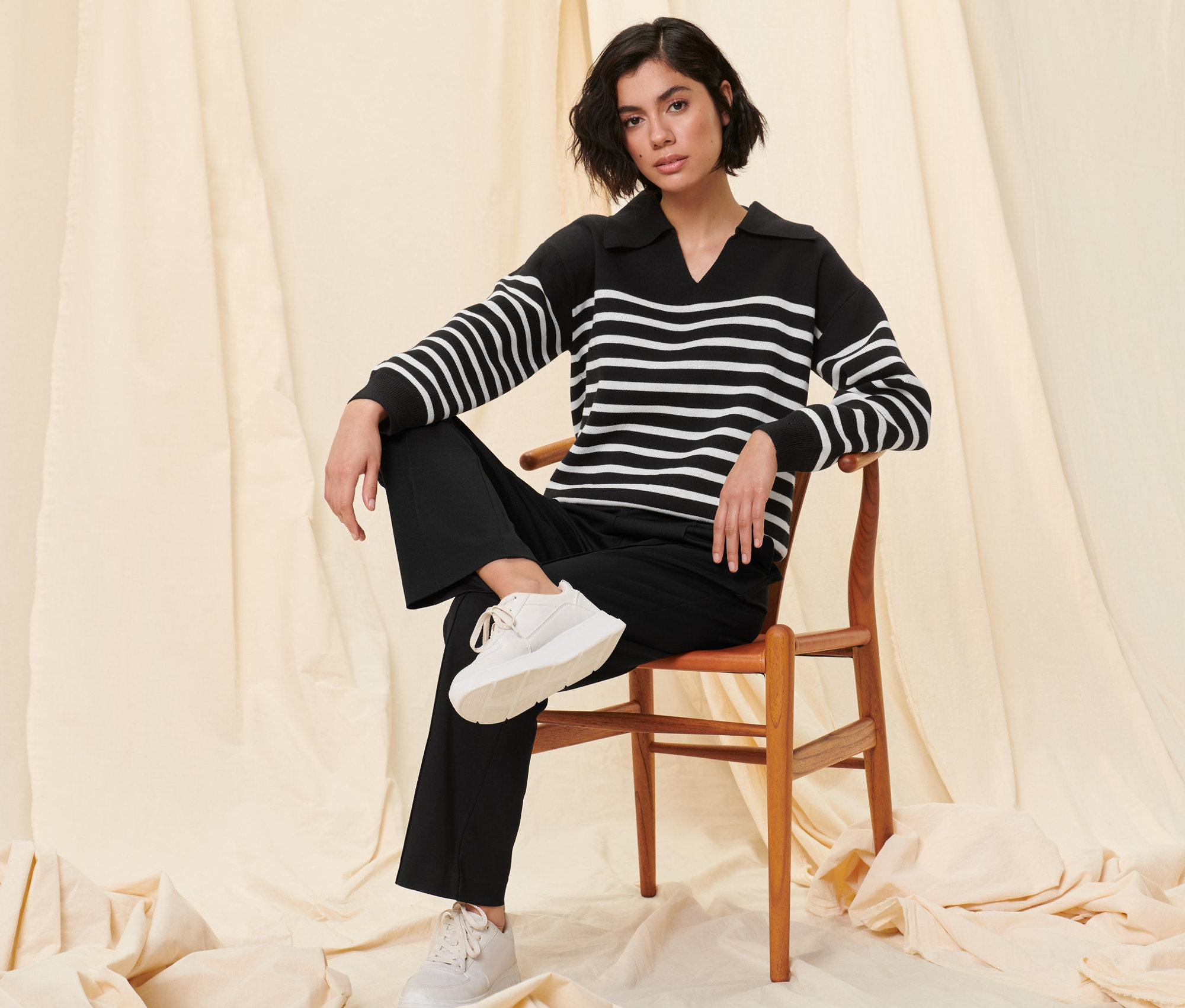 Nő ül egy fa széken, csíkos kötött pulóvert és fekete szabadidőnadrágot visel.