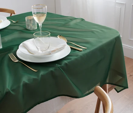 Egy zöld organza asztalterítő, kb. 140 x 180 cm, egy asztalon terül el, melyen fehér tányérok, poharak és arany evőeszközök találhatók.
