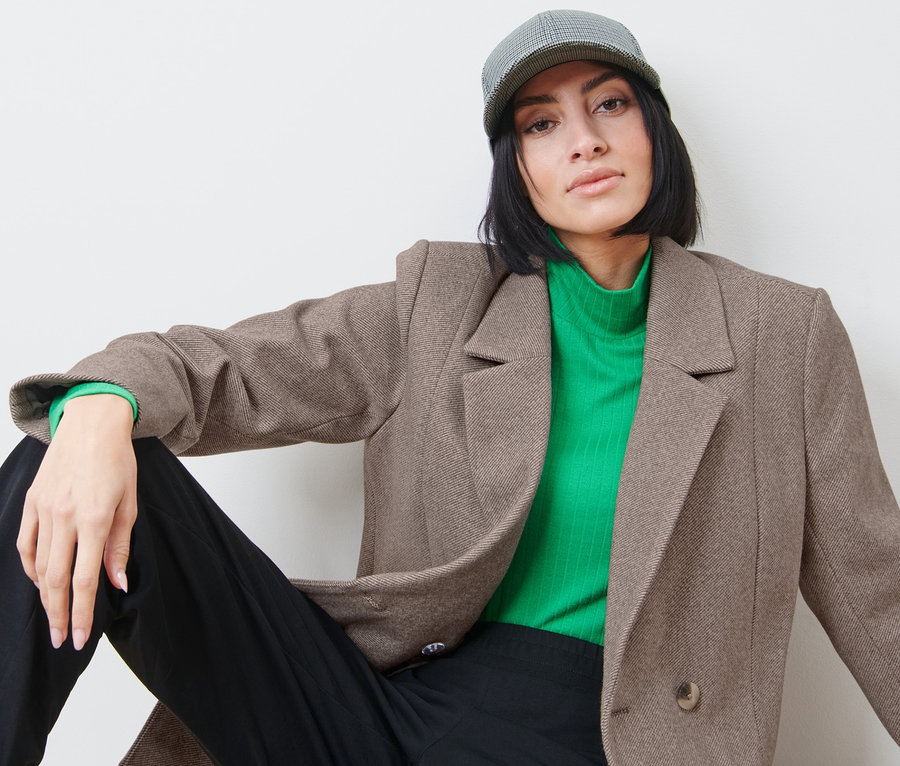 Nő ül, és blézert, zöld garbót és baseballsapkát visel.