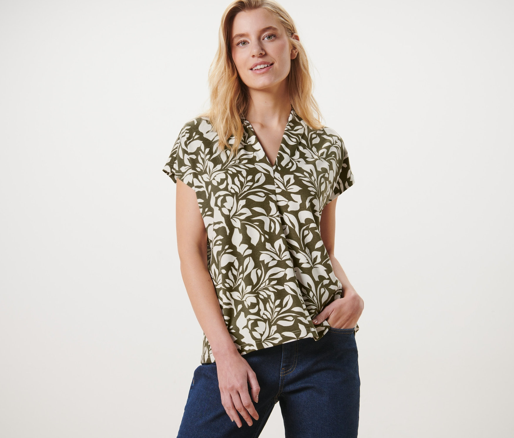 Nő pózol mintás pólóblúzban V-nyakkivágással és farmerban.