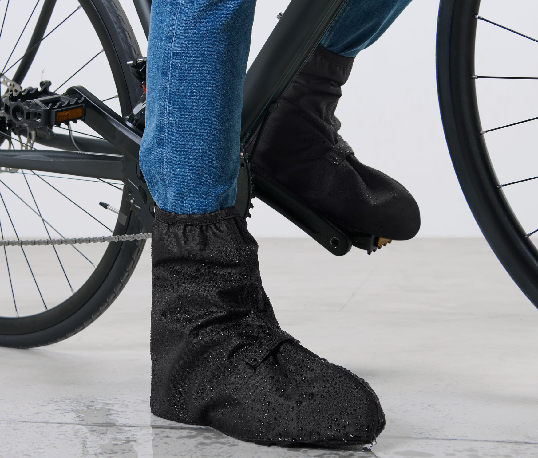 Egy kerékpáros lábai kék farmerben és fekete kamásli, vízálló cipőben egy kerékpáron.