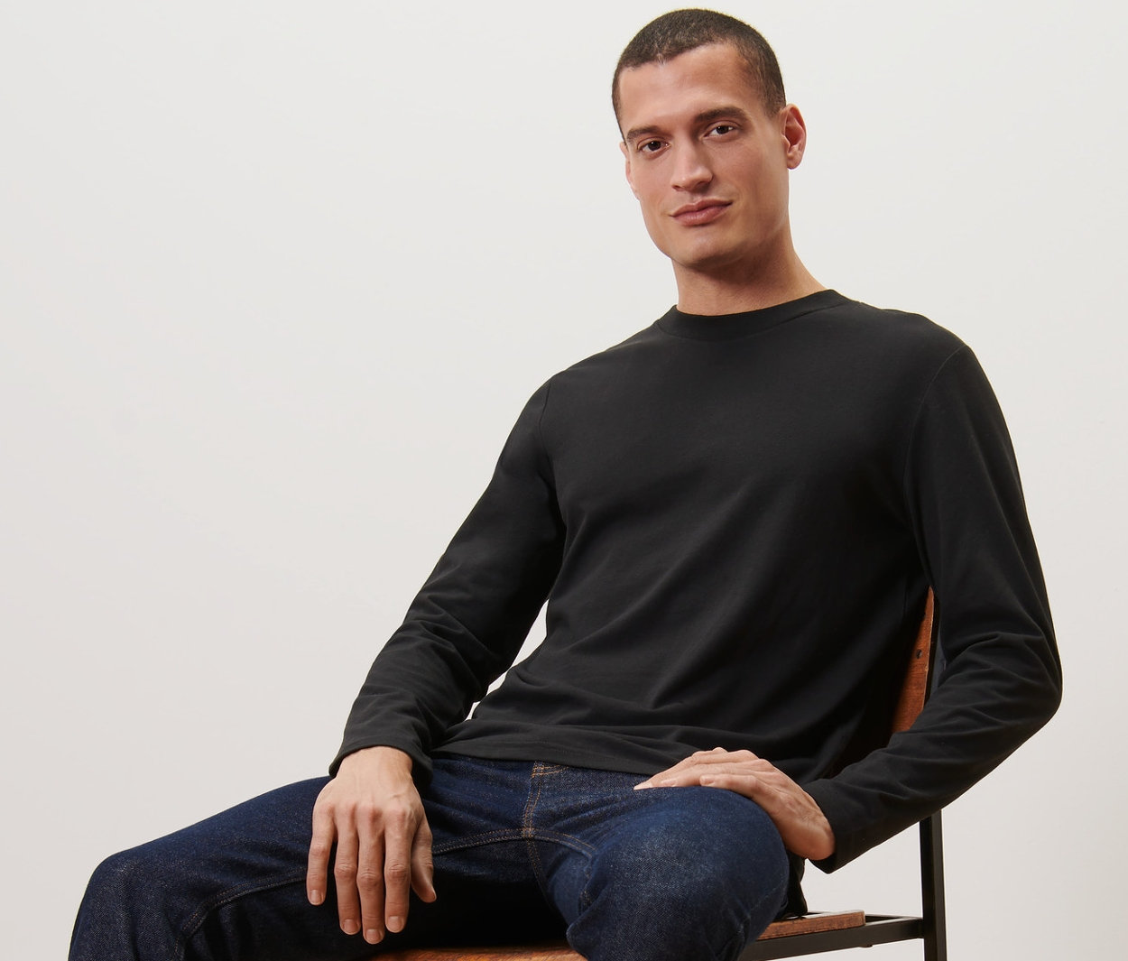 Férfi ül egy széken, fekete 2 férfi hosszú ujjú felsőt, farmert és edzőcipőt visel.