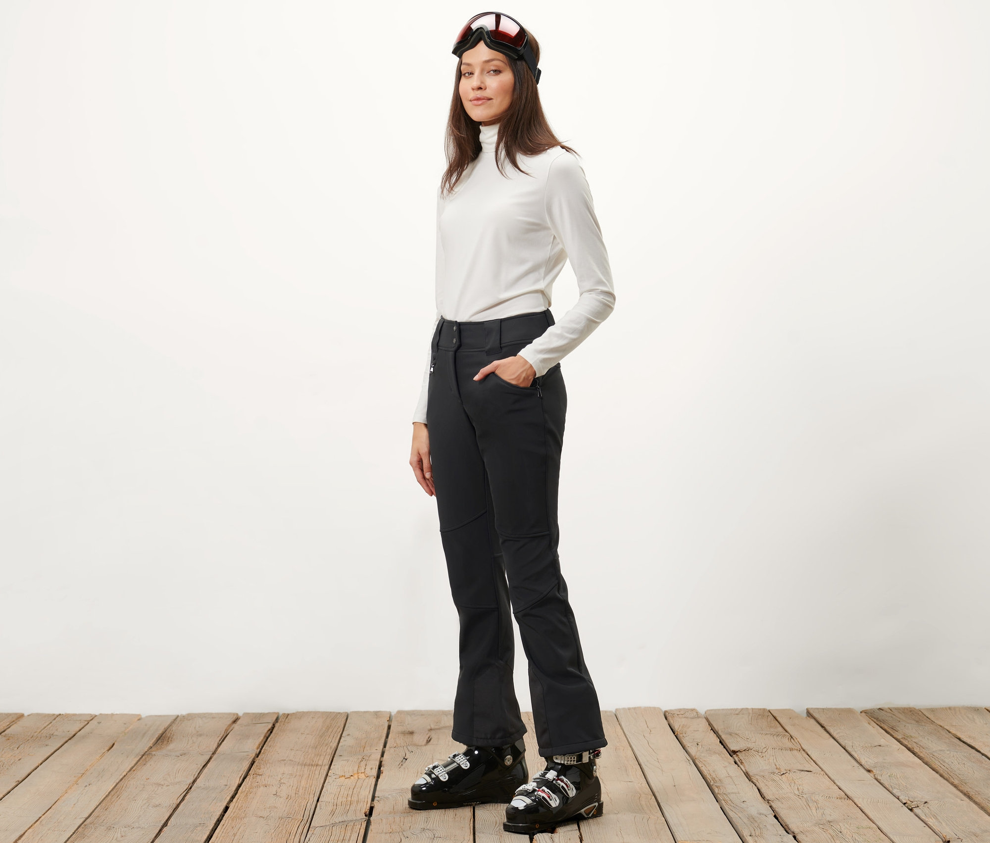 Nő pózol fekete softshell jet sínadrágban és síbakancsban.