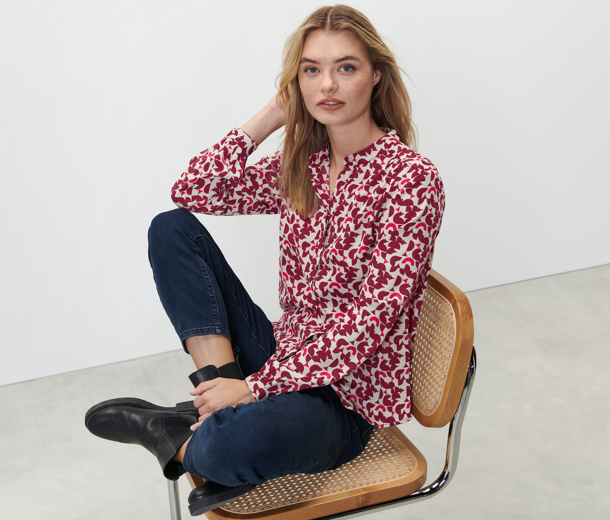 Nő ül egy széken, és egy piros/krém mintás női hosszú ujjú blúzt visel.