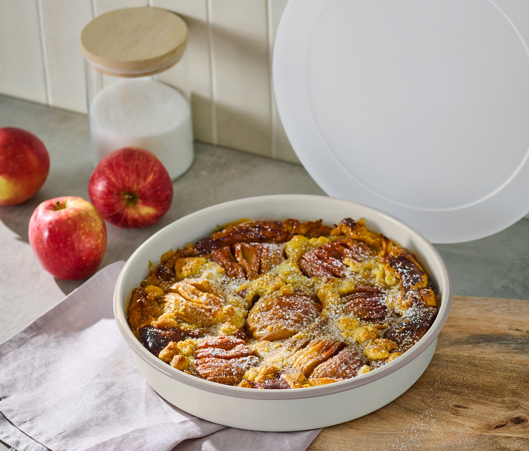 Almás pite zománcozott piteformában, fa deszkán, almák és cukortartó mellett.