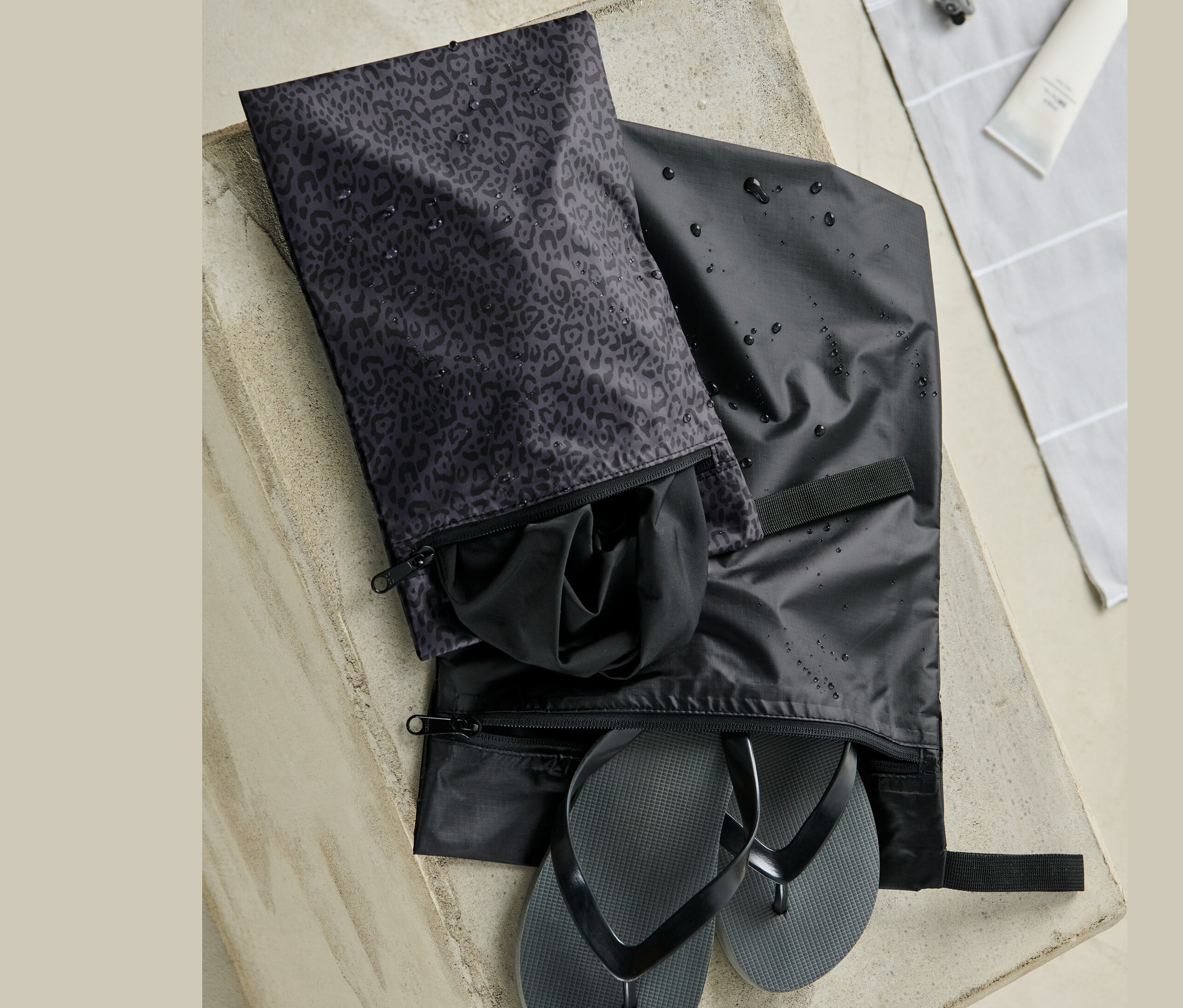 Két vízálló tárolótasak egy betonfelületen. Az egyikben fekete flip-flop papucsok, a másikban fekete ruhák vannak.
