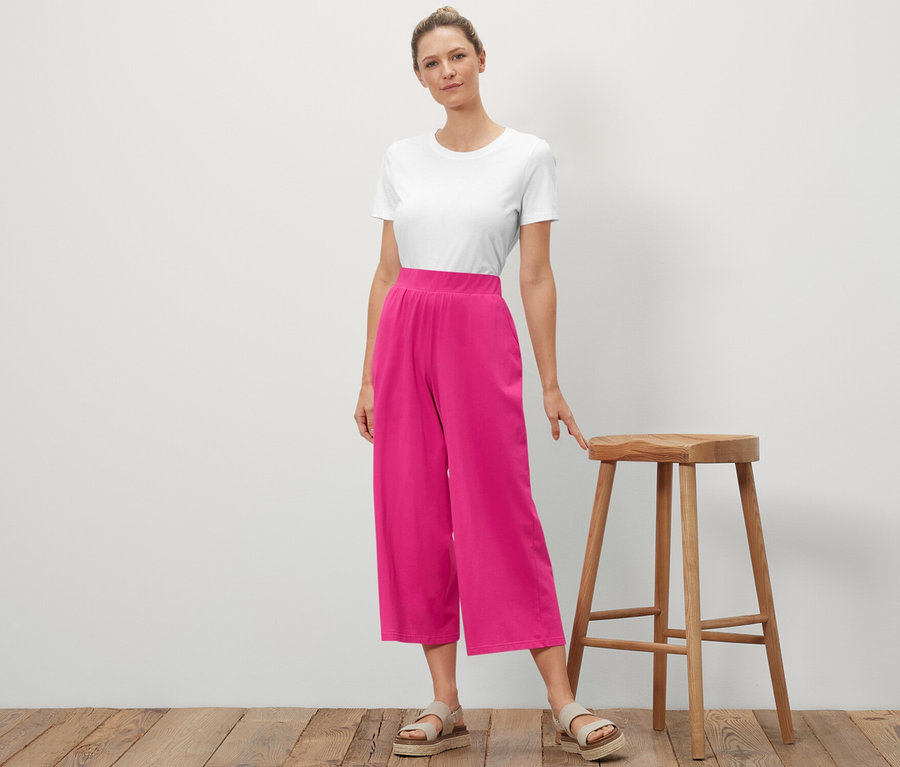 Egy nő áll egy fa szék mellett és pózol. Fehér pólót és rózsaszín bő szárú jerseynadrágot visel.