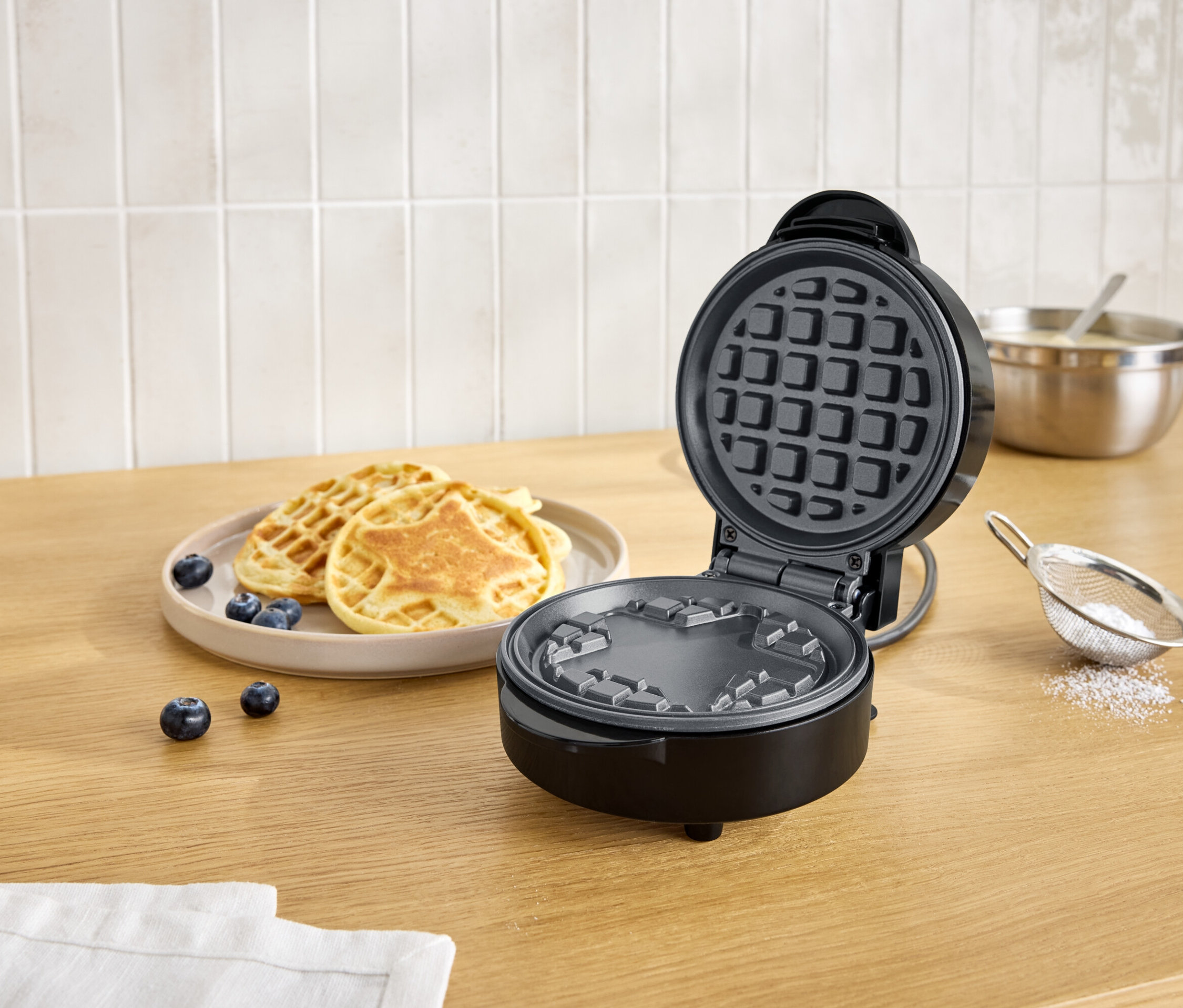 Nyitott, fekete gofrisütő egy tányér gofri és áfonya mellett egy fa asztalon.