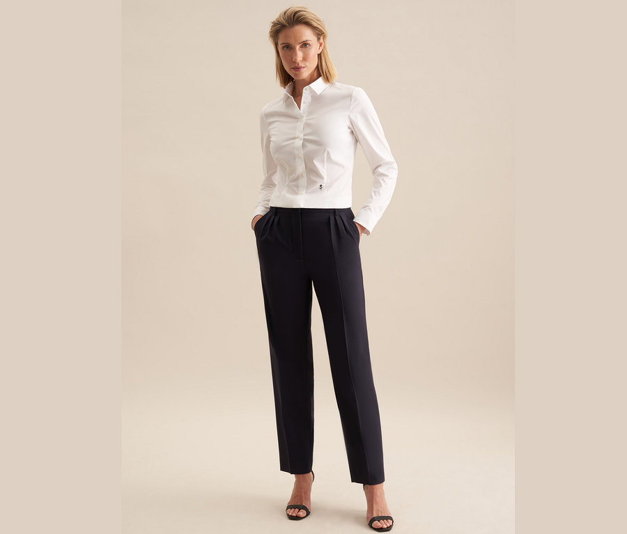 Egy nő áll, kezével a zsebében. Fehér Seidensticker slimfit blúzt, sötétkék nadrágot és fekete szandált visel.