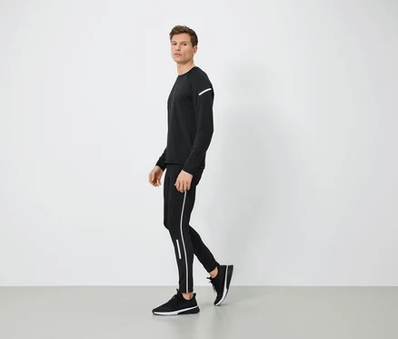 Férfi fekete sportruházatban, fényvisszaverő részletekkel sétál szürke padlón.