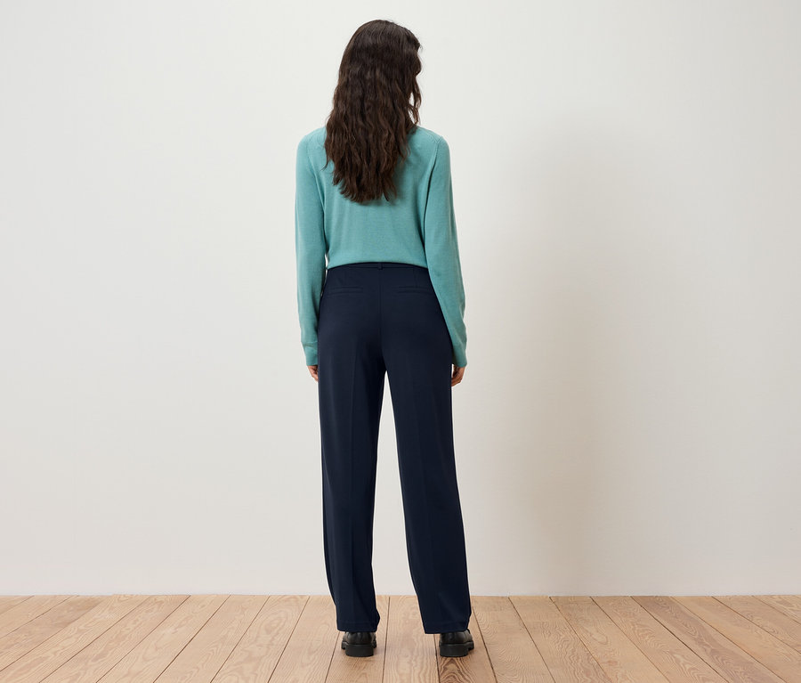 Hátulról látható egy sötétbarna hajú nő, aki türkiz pulóvert és sötétkék nadrágot visel.