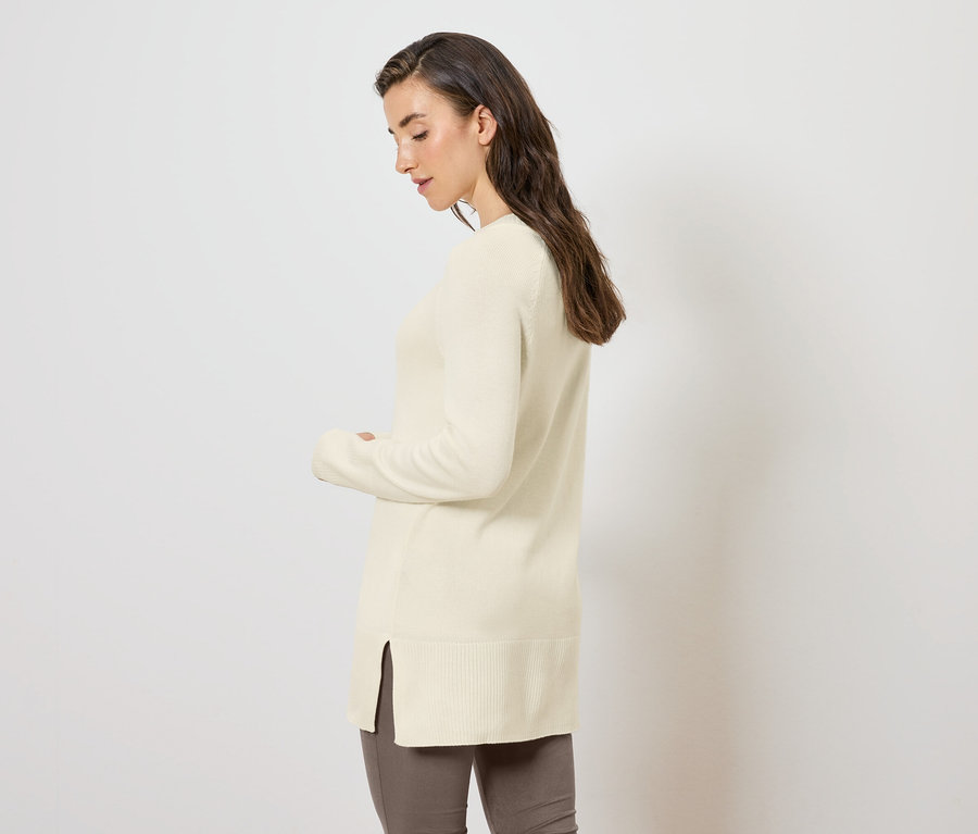 Nő oldalra pózol egy hosszú, krémszínű kötött pulóverben.