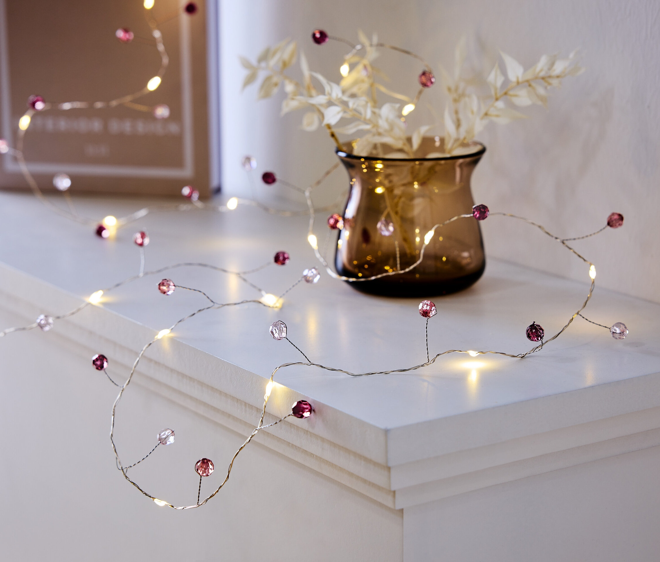 Dekoratív fényfüzér lila és átlátszó gyöngyökkel egy fehér polcon, egy vázával és egy könyvvel.