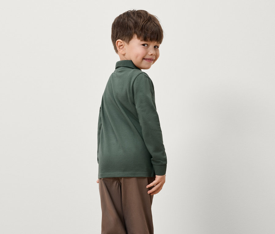 Egy fiú visszanéz a válla fölött, zöld, hosszú ujjú pólót és barna nadrágot visel.