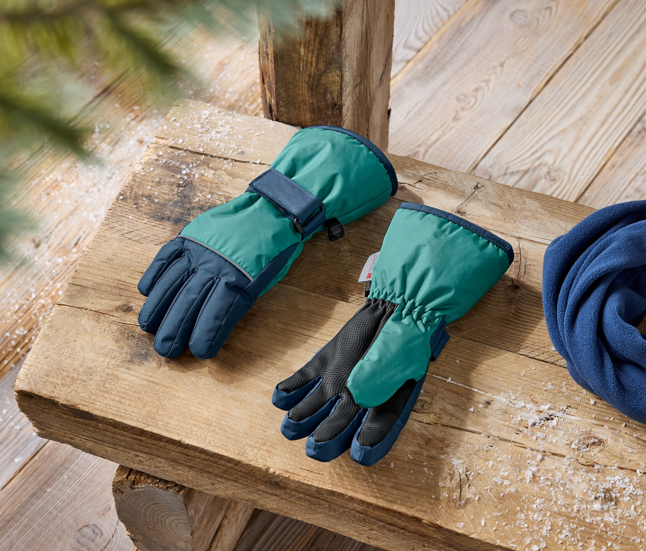 Zöld és sötétkék téli kesztyűk és egy kék polár sál fekszik egy fa padon.