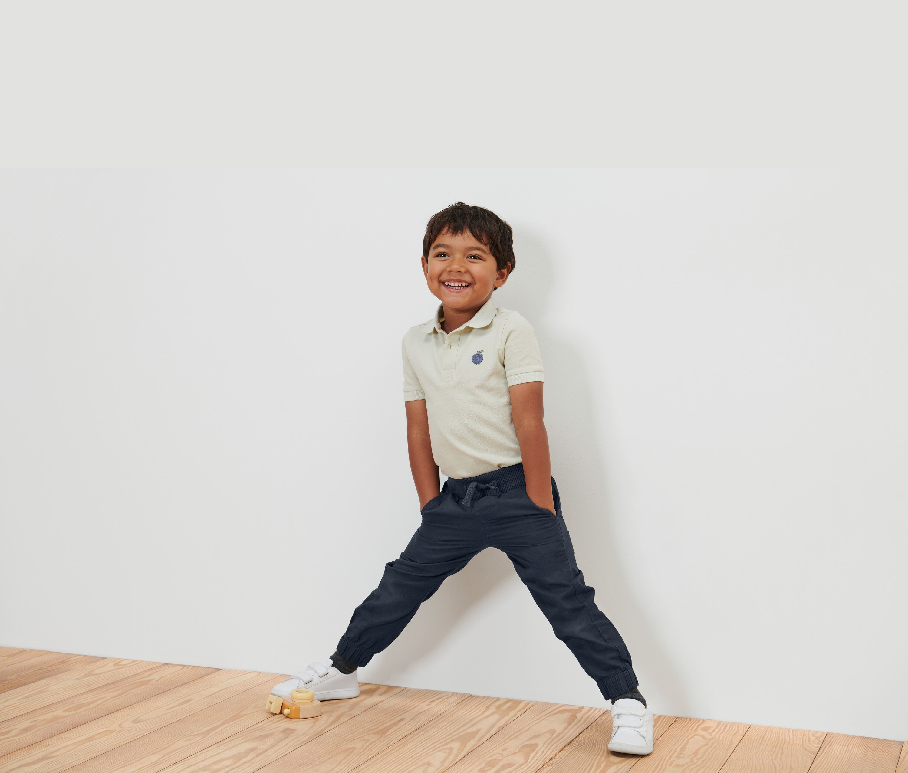 Egy mosolygó fiú bézs színű pólóban, sötétkék nadrágban és fehér cipőben áll egy fehér falnál.
