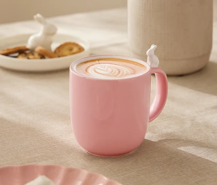 Az asztalon egy rózsaszín bögre áll, fehér nyuszi díszítéssel, tele latte artos itallal. A háttérben egy fehér váza virágokkal, egy tányér süteménnyel és egy nyuszi figura látható.