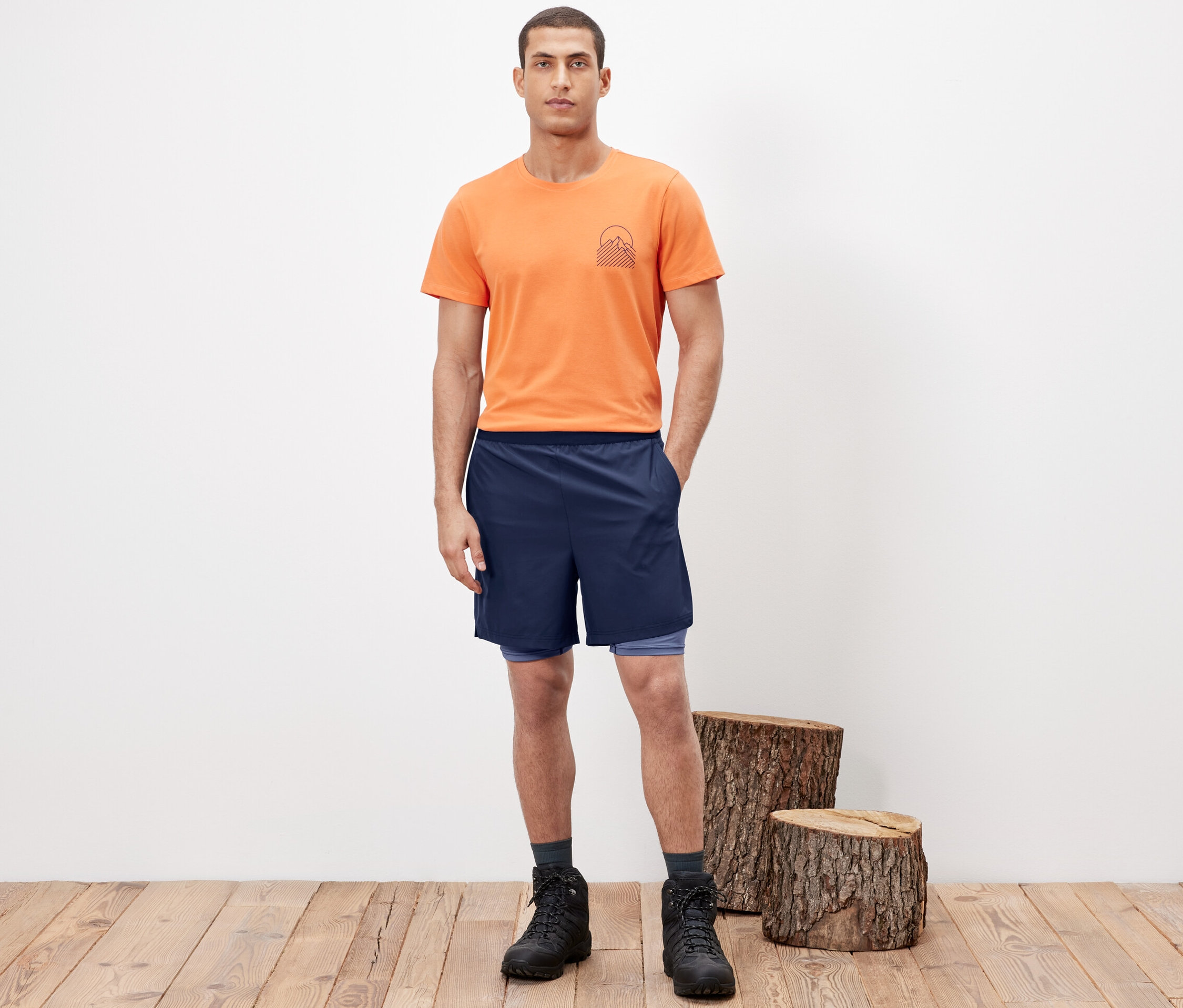 Férfi pózol 2 az 1-ben funkcionális rövidnadrágban, narancssárga pólóban és túrabakancsban.