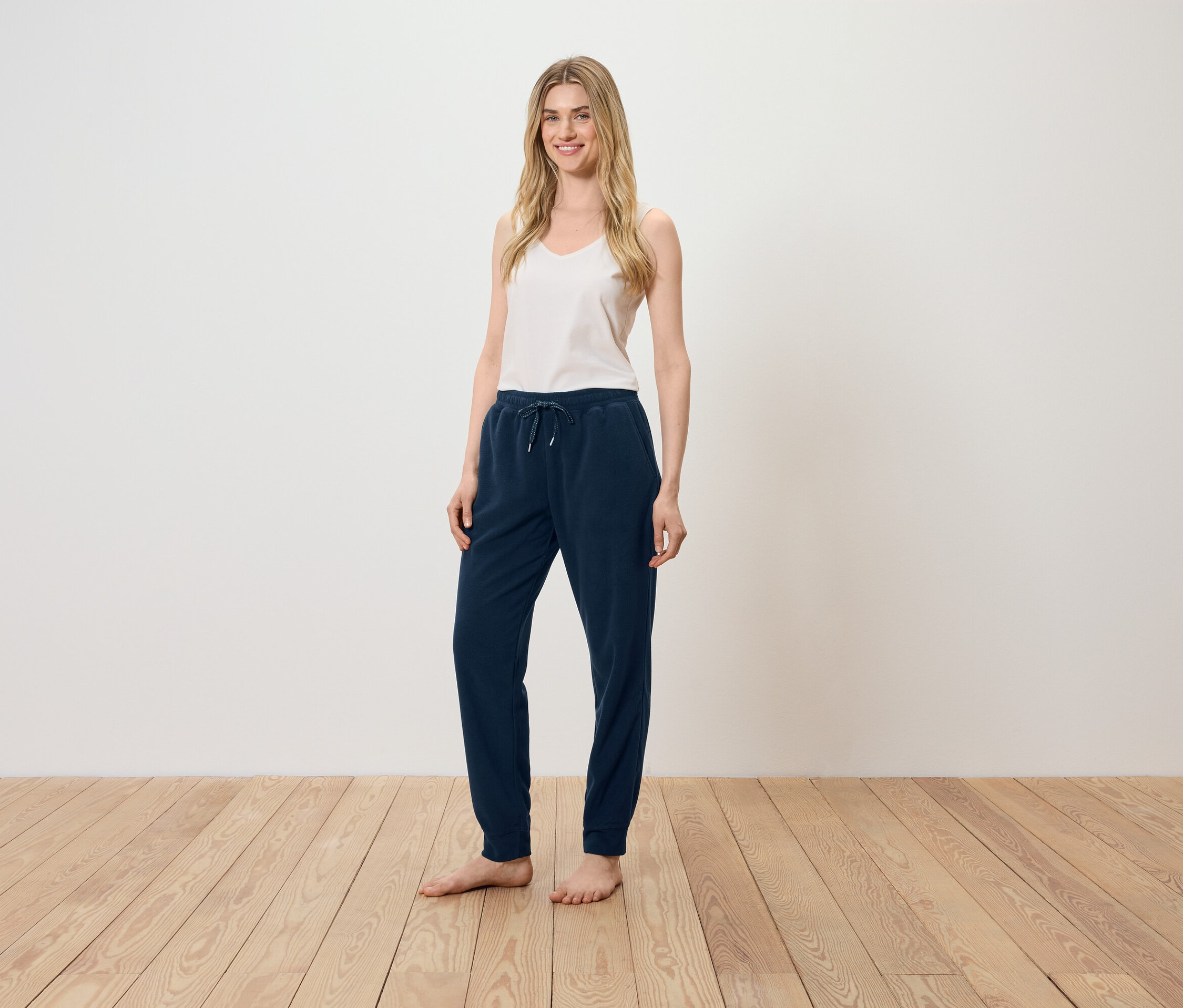 Egy nő áll a fapadlón, fehér felsőt és sötétkék melegítőnadrágot visel.