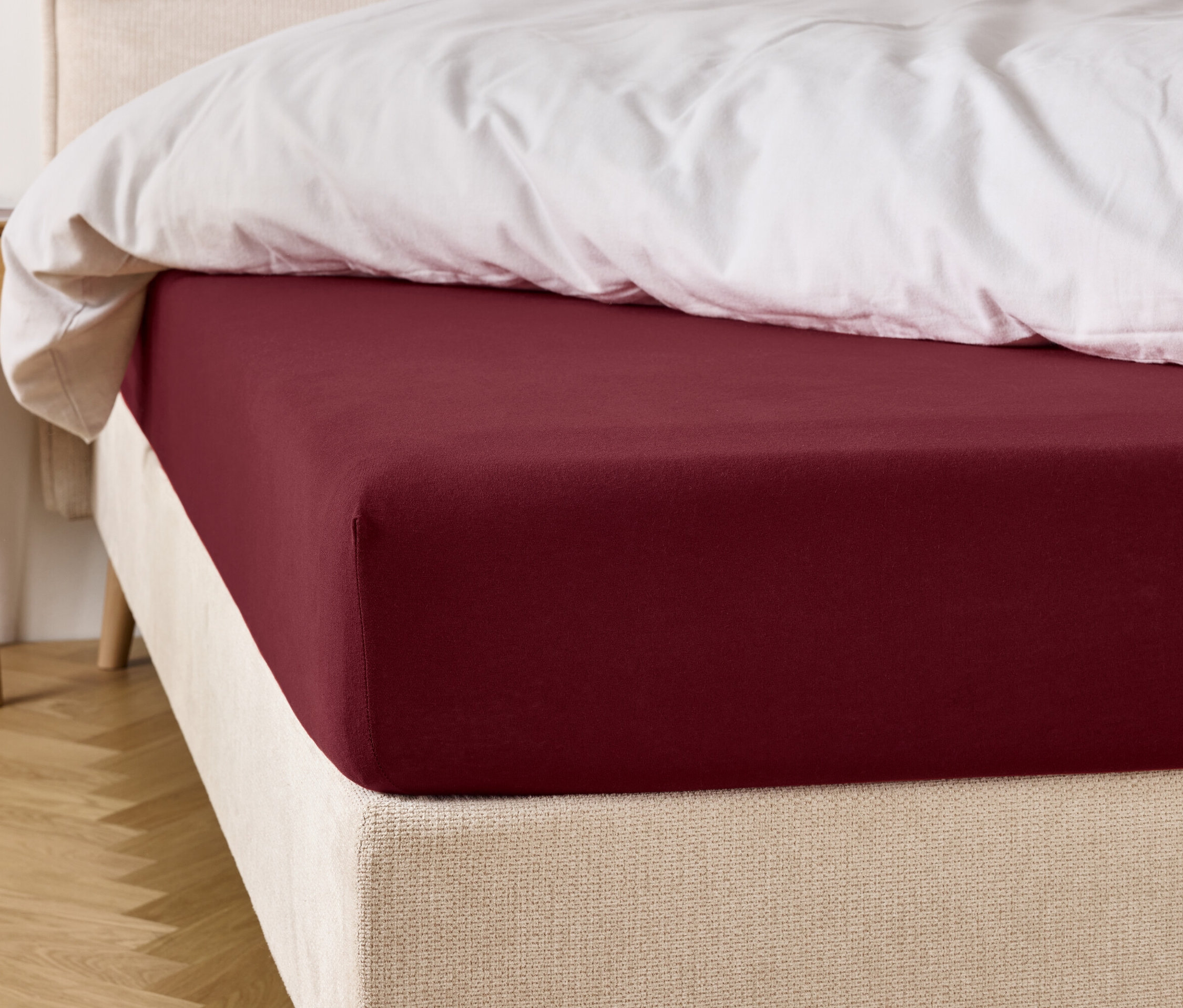 Bordó színű gumis lepedő egy ágyon, fehér paplannal.