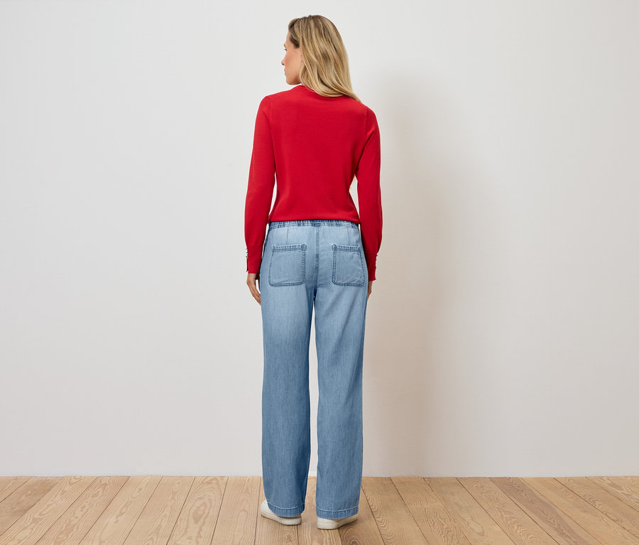 Egy nő áll háttal a kamerának, piros pulóvert és kék farmert visel.
