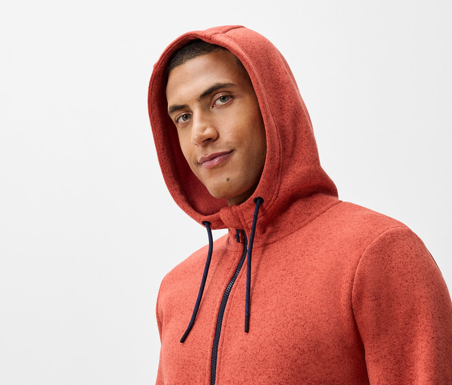 Egy férfi portréja, narancssárga pulóverben, kapucnival és sötétkék cipzárral.