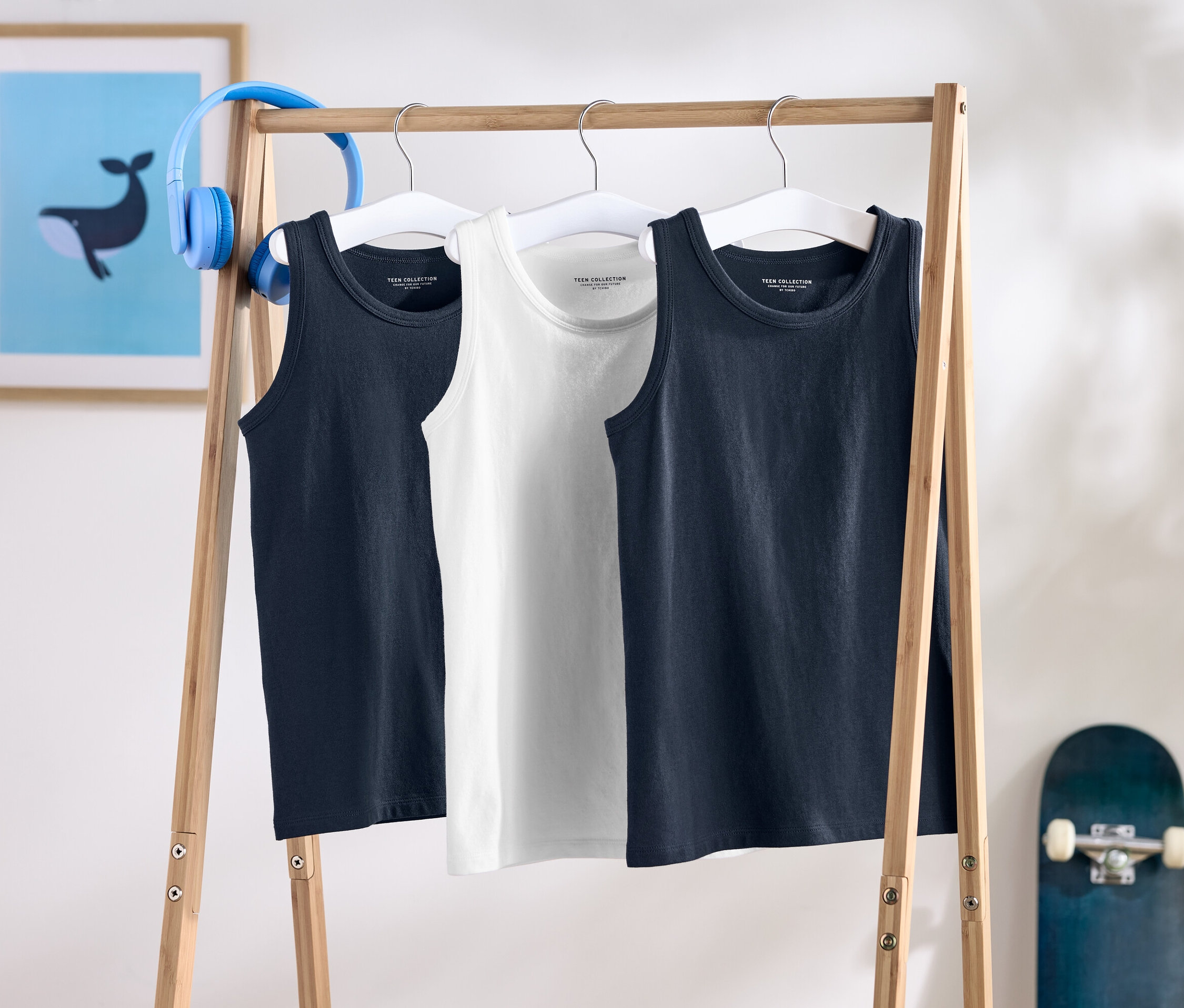 Három ujjatlan trikó lóg egy fa állványon, mögöttük gördeszka és fejhallgató.