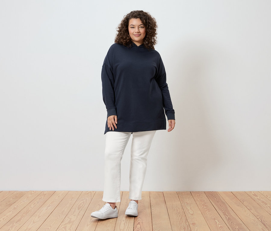 Egy göndör hajú nő áll egy fal előtt, tengerészkék kapucnis pulóvert, fehér farmert és fehér tornacipőt visel.