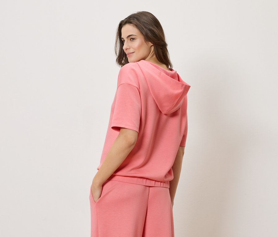 Nő korall színű, rövid ujjú kapucnis pulóvert és hozzáillő korall színű rövidnadrágot visel.