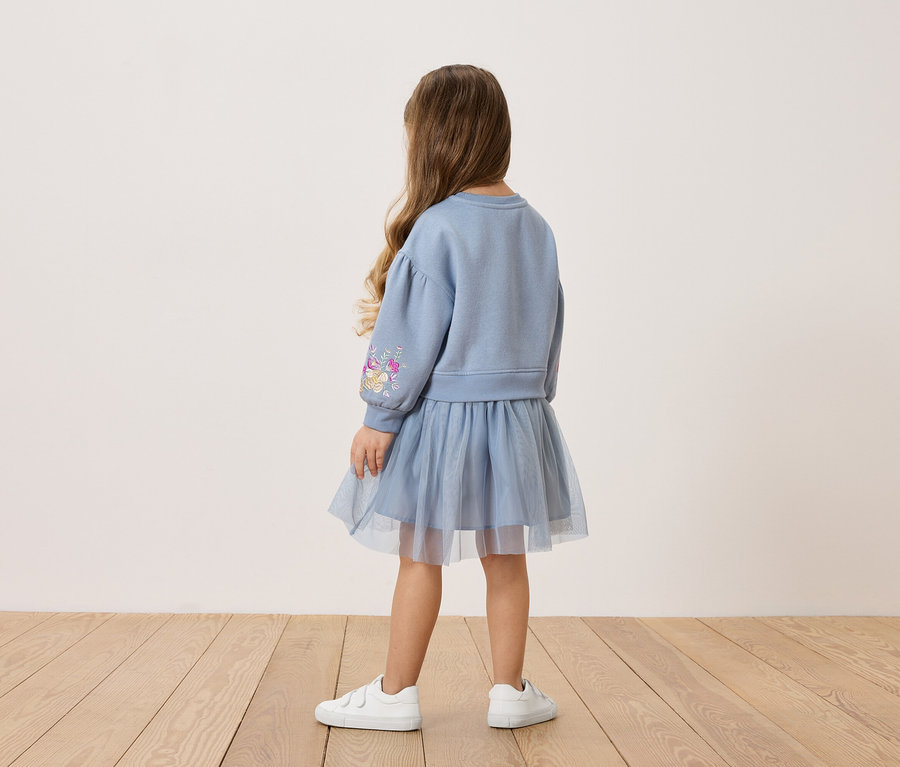Hátulnézet egy kislányról, hosszú barna hajjal, kék pulóverben, virágmintával az ujján, hozzáillő tüllszoknyával és fehér tornacipővel.