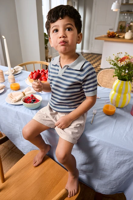 Fiú ül az asztal szélén, villával epret eszik, Kinder-Shorts és Kinder-Strick-Poloshirt van rajta. Az asztalon váza és tortatálca.